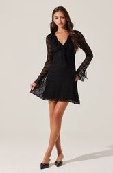 A woman is wearing a black lace dress with long, semi-sheer, striped sleeves. The dress has a V-shaped neckline with ruffle detailing along the collar and down the front. The hem of the dress falls above the knees. She is also wearing black pointed-toe shoes with a bow detail on the front.